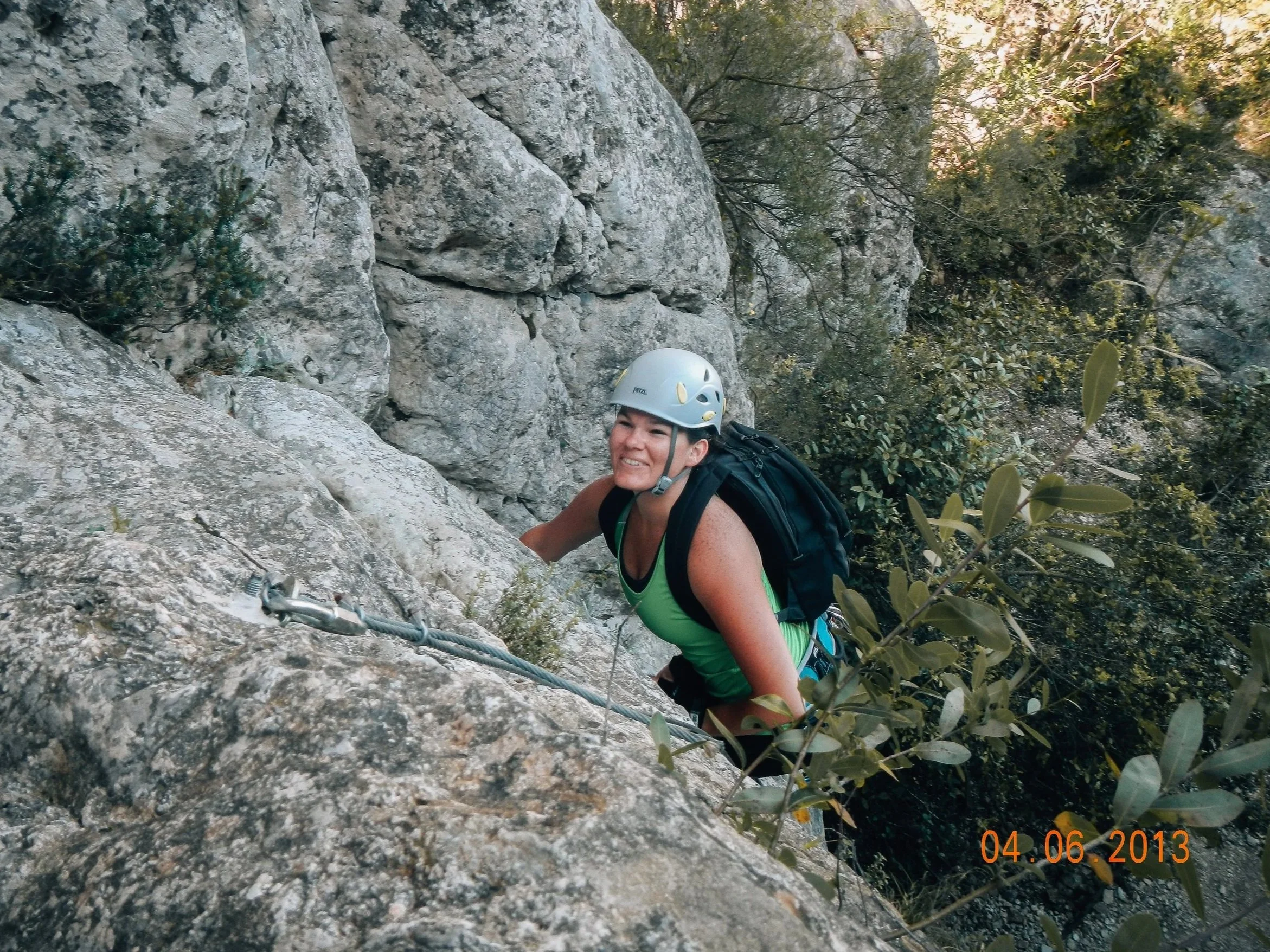 Woman with long brown hair, climbing the mountains of tivissa, doing a via ferrata, with a black bag on and a Petzl helmet on.