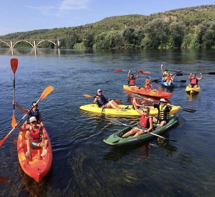 Group of people kayaking and paddleboarding on a calm river, surrounded by green hills, with a bridge in the background.