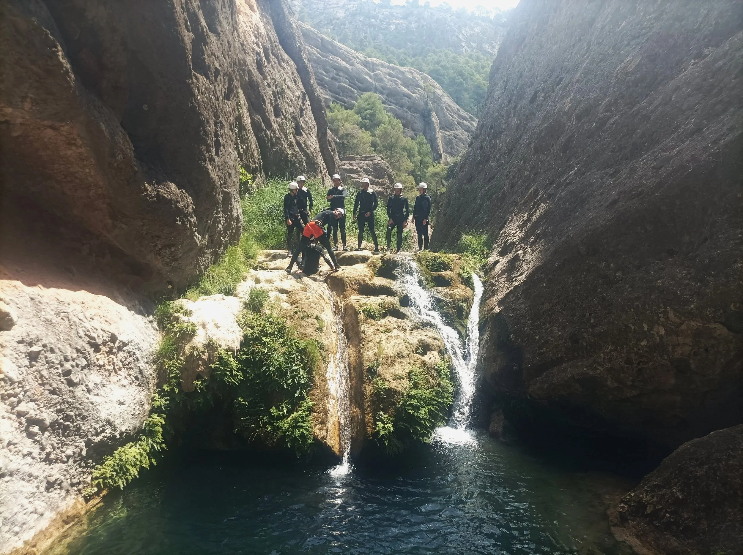 Group of people exploring a rocky waterfall in a canyon with water flowing over rocks surrounded by cliffs and greenery.