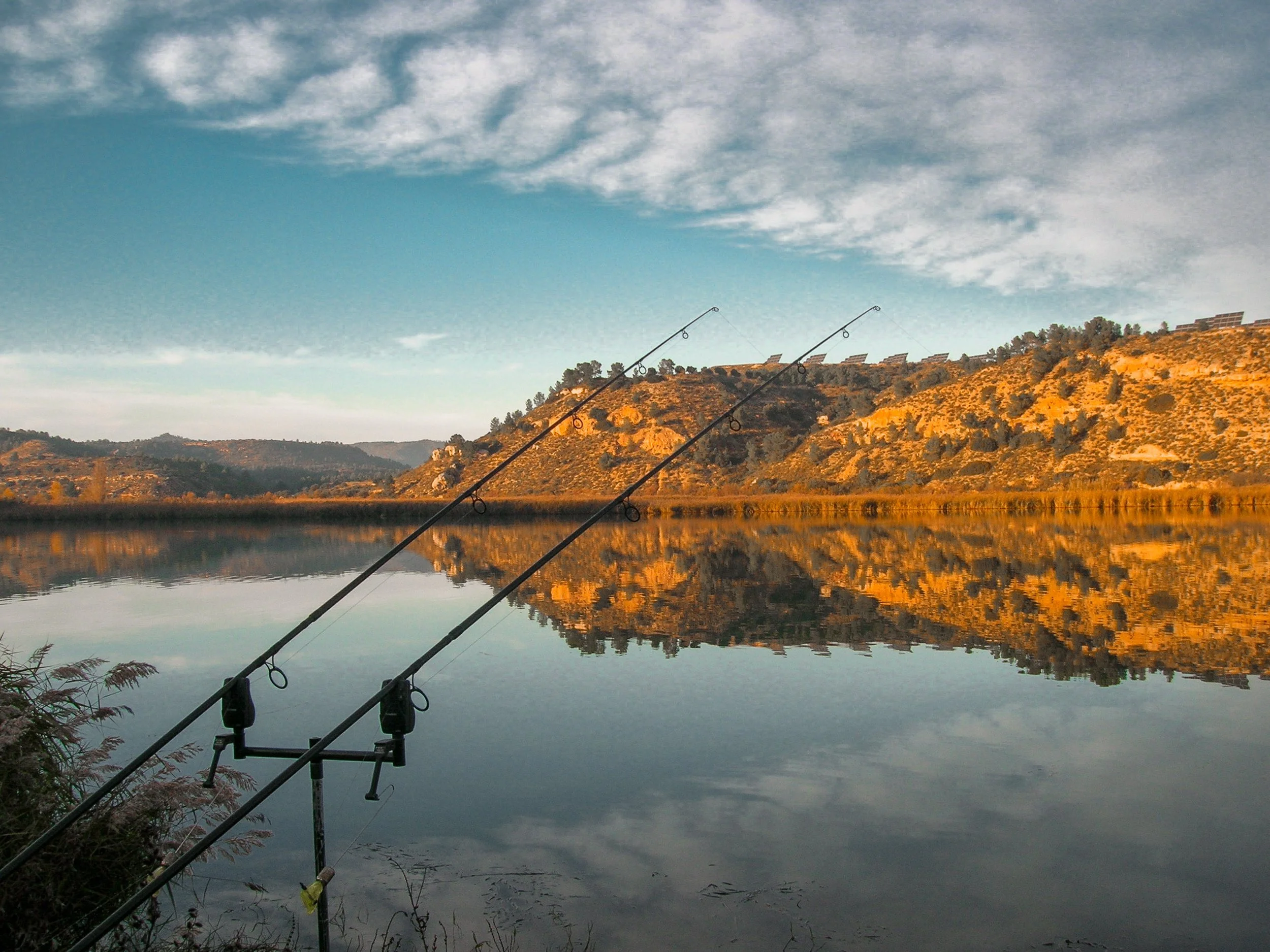 Two fishing rods set up by a calm lake with mountains and sky in the background, reflected in the water.