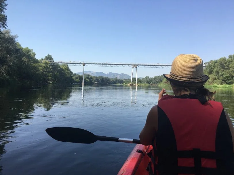 Person in a kayak on a river, wearing a straw hat and red life jacket, pointing toward a bridge in the distance with mountains and trees on either side.