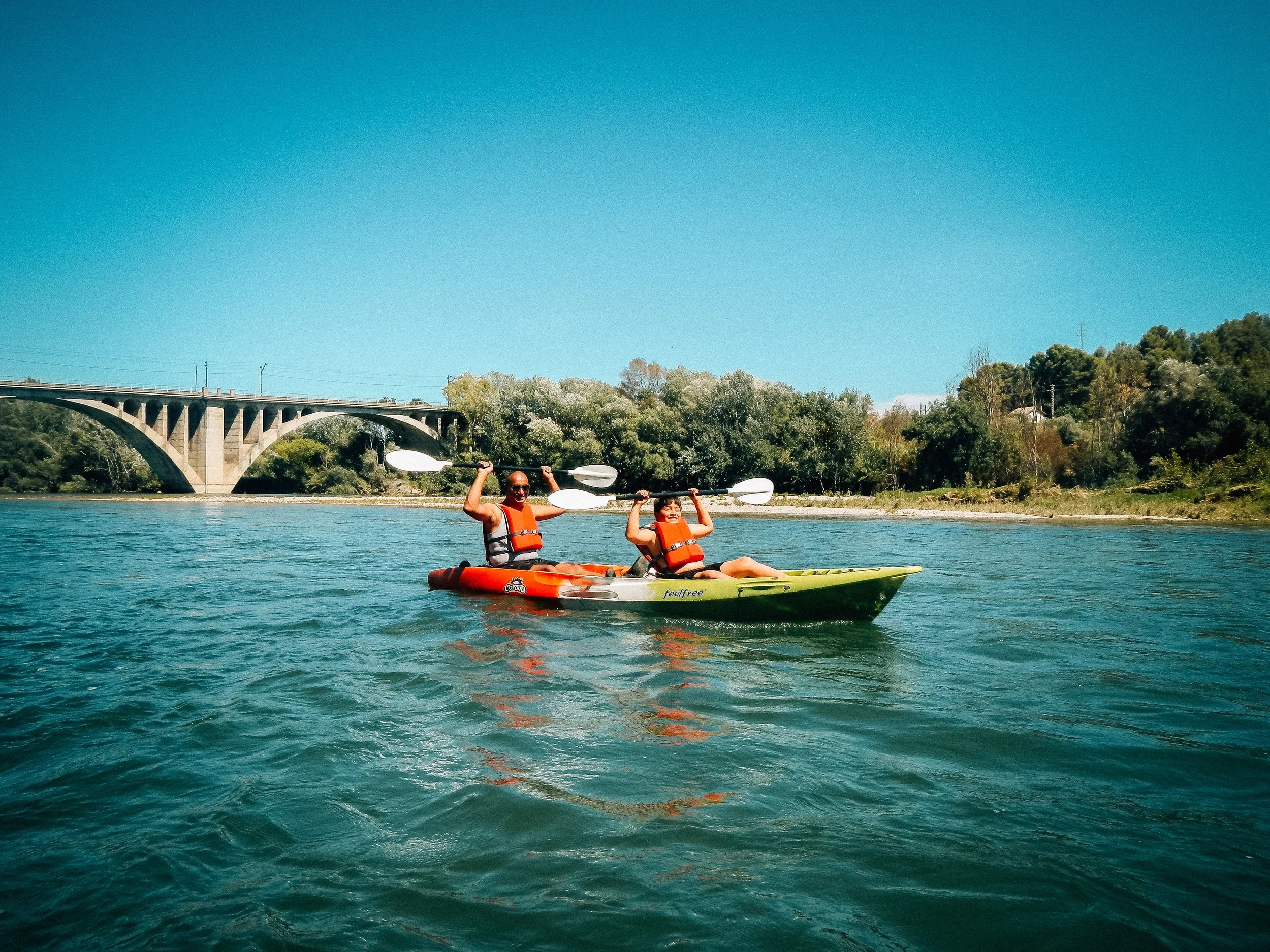 Two people kayaking on a river with trees and a bridge in the background under a clear blue sky.