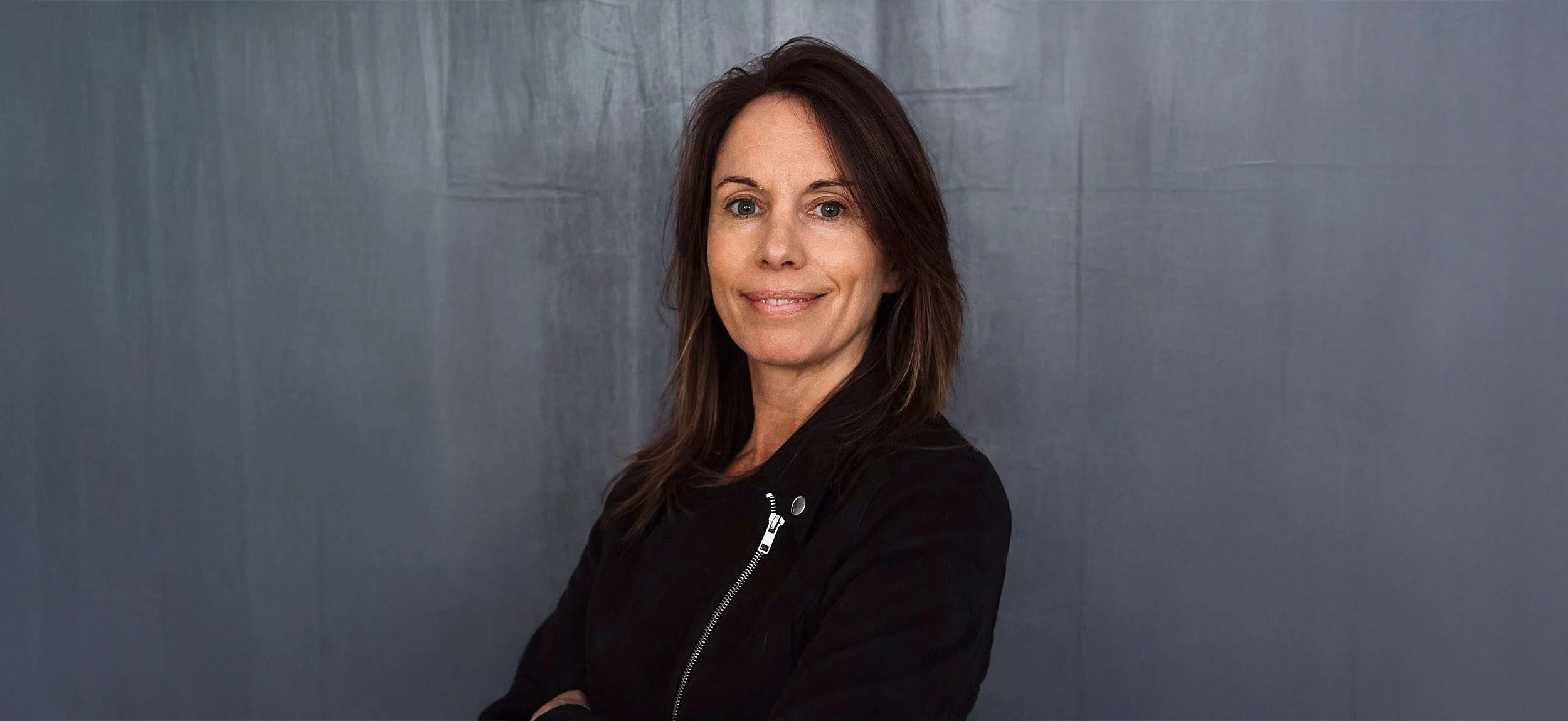 En kvinna med brunt hår, svart jacka, står framför en grå vägg, ler och tittar mot kameran.
