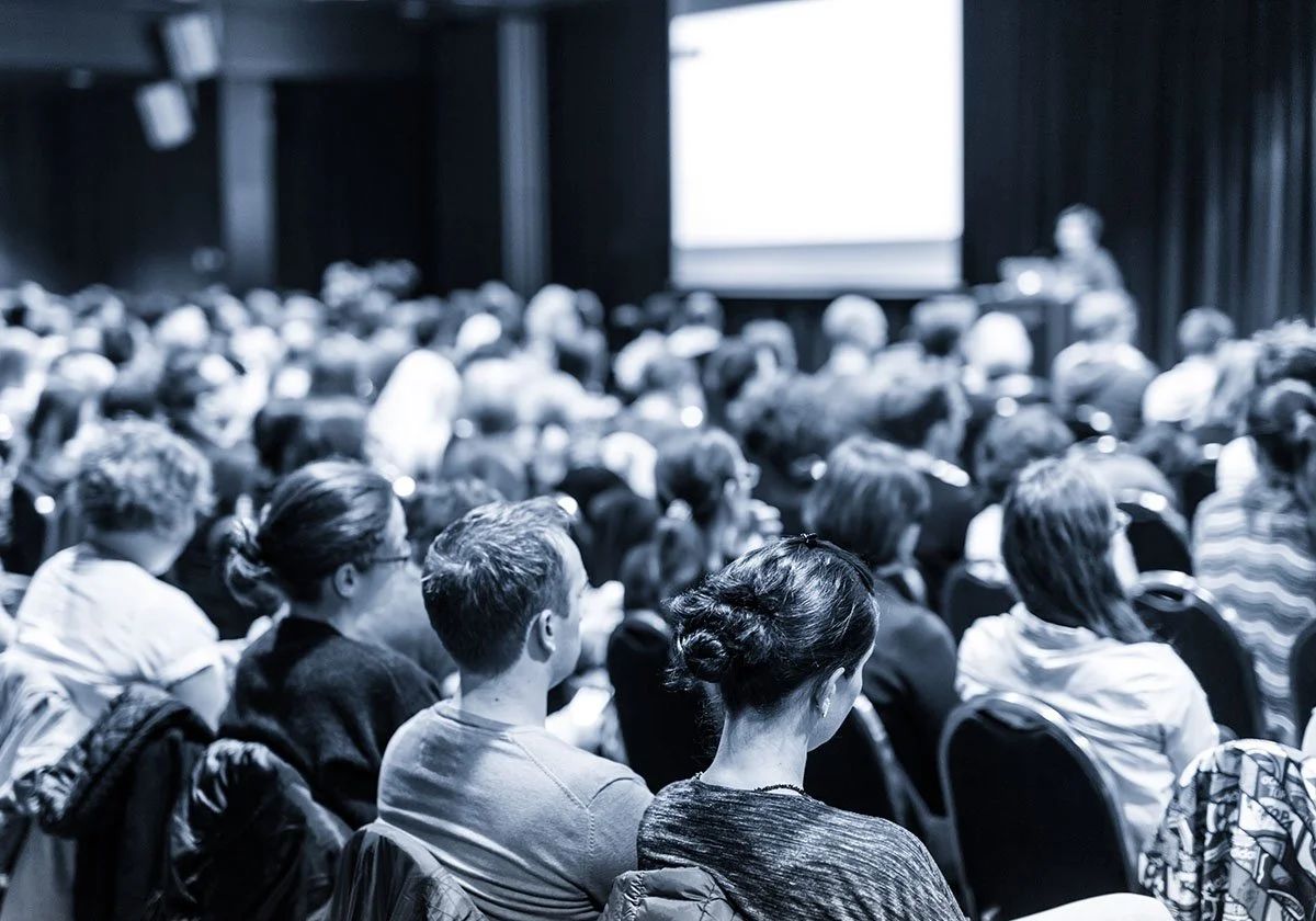 En stor publik av människor sitter i ett mörklagt auditorium och tittar mot en konferens eller presentation som visar en tom whiteboard eller skärm.