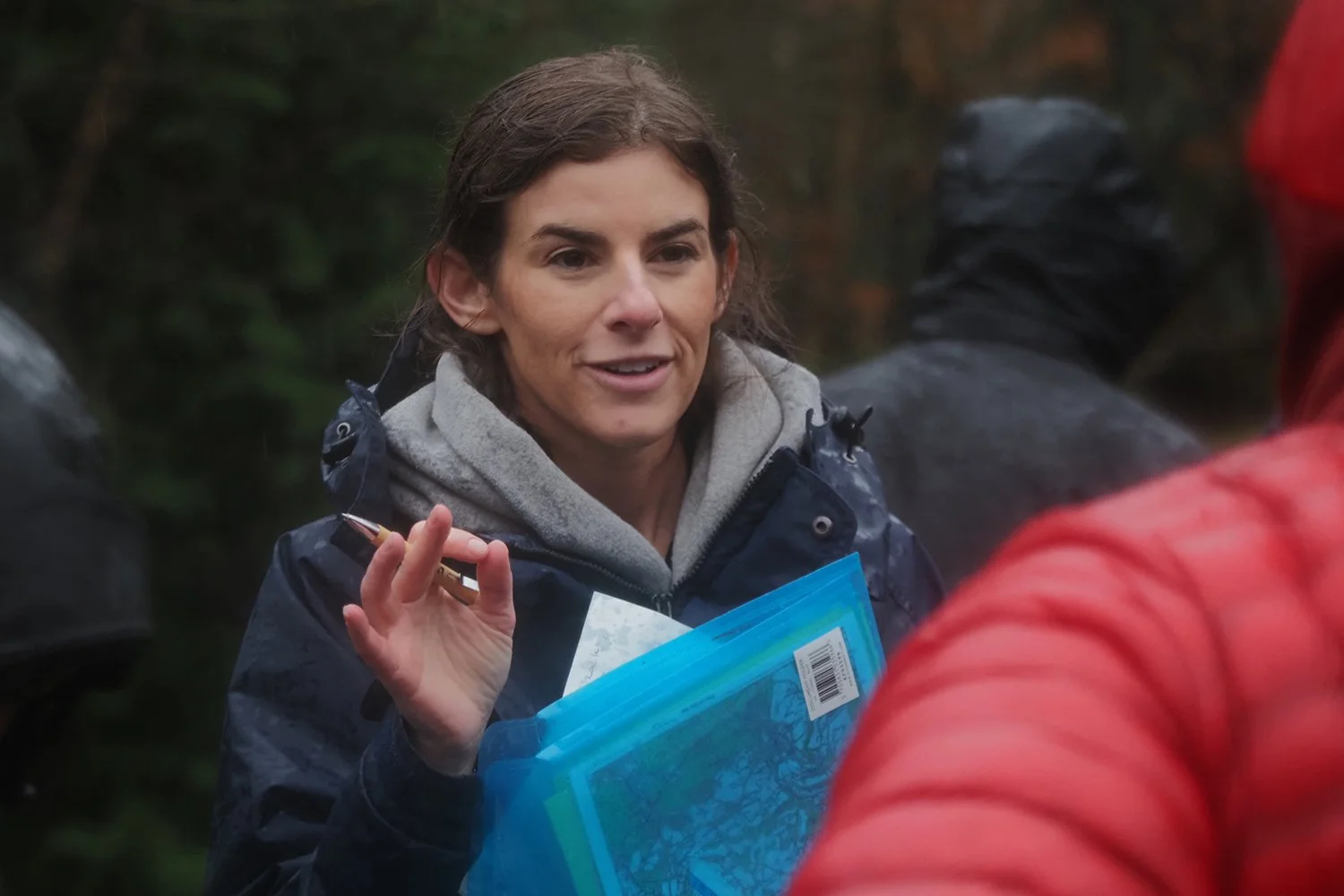 A woman with brown hair, wearing a dark jacket, gray hoodie, and holding a blue map, is talking with a person in a red jacket outdoors in a wooded area.