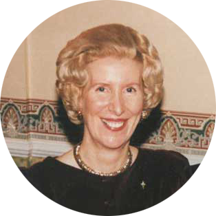 A woman with blonde curly hair, wearing a black outfit and pearl necklace, smiling indoors in front of a beige wall with a decorative border.
