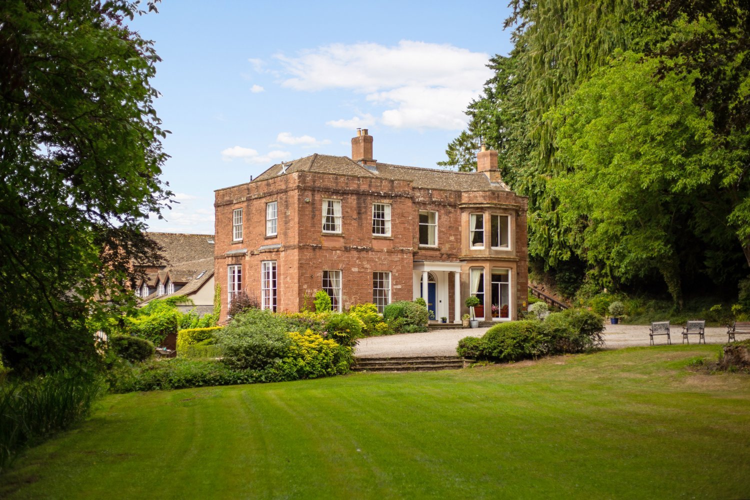 A large, historic red-brick house surrounded by a well-maintained garden with green lawns, shrubs, and trees on a clear day with a blue sky and some clouds.