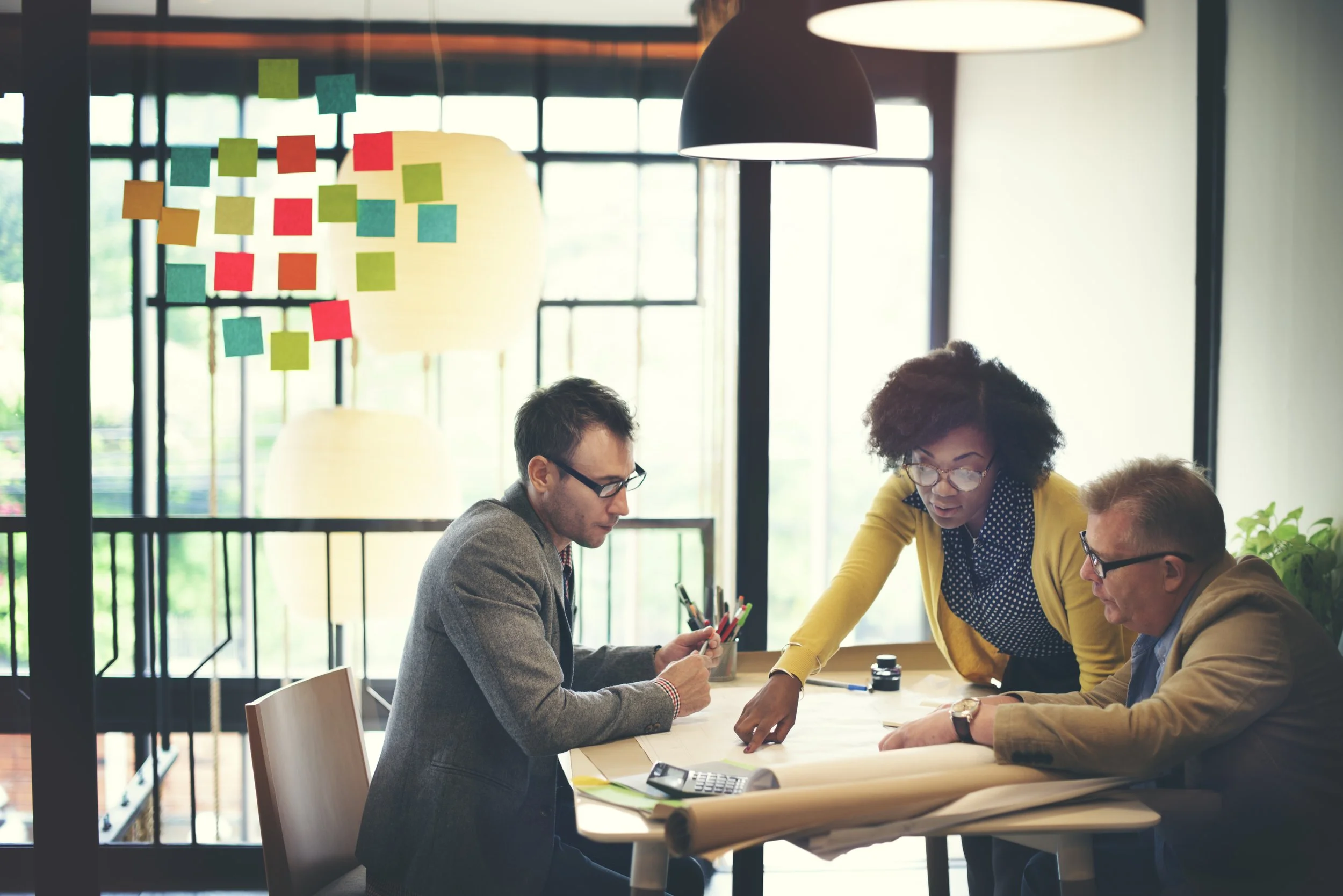 Three business professionals working together at a table in a bright office space with large windows and a modern industrial design. The group is reviewing documents and using office supplies.