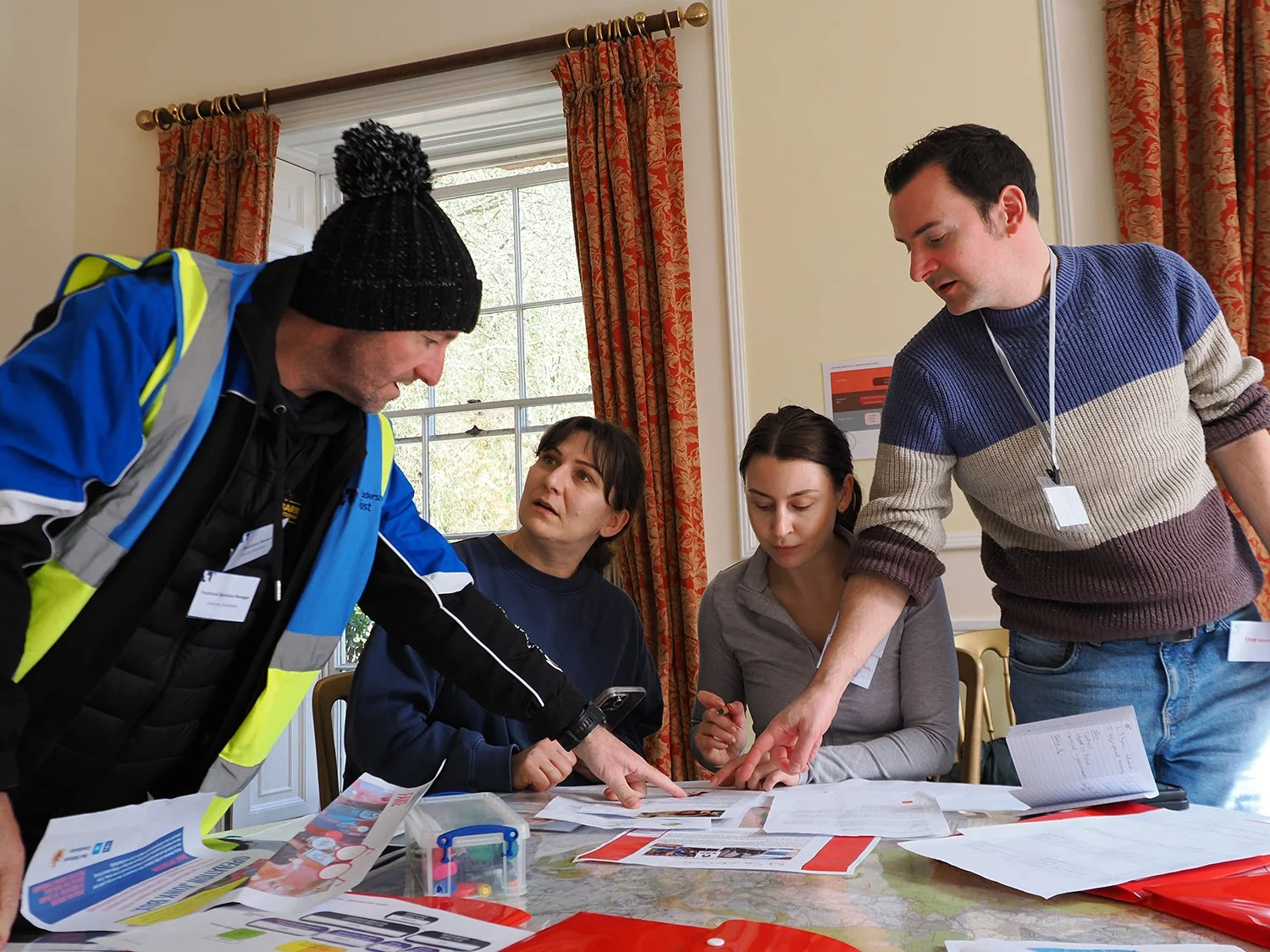 Four people gathered around a table examining documents and papers, with a man in a blue and yellow jacket pointing at something.