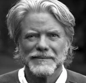 A black-and-white close-up portrait of an older man with gray hair, a beard, and a mustache, looking directly at the camera with a neutral expression.