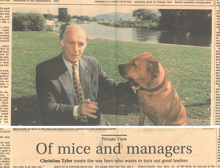 A newspaper clipping showing a man in a gray suit sitting outdoors next to a large brown dog. The man appears to be touching the dog, with a river and trees in the background.