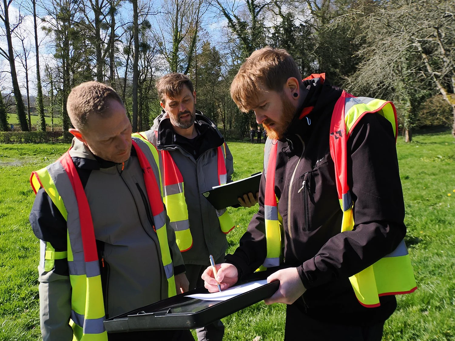 Three men wearing high visibility vests standing outdoors on a grassy field, reviewing documents or a map.