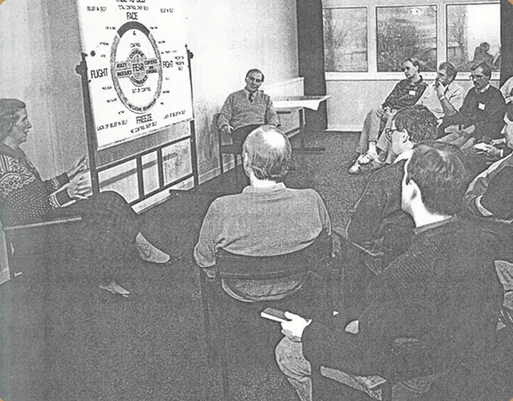 A black-and-white photo of a group of people attending a lecture or presentation in a conference room. A woman is speaking in front of a whiteboard with diagrams and text, while the audience, seated in chairs, listens and takes notes.