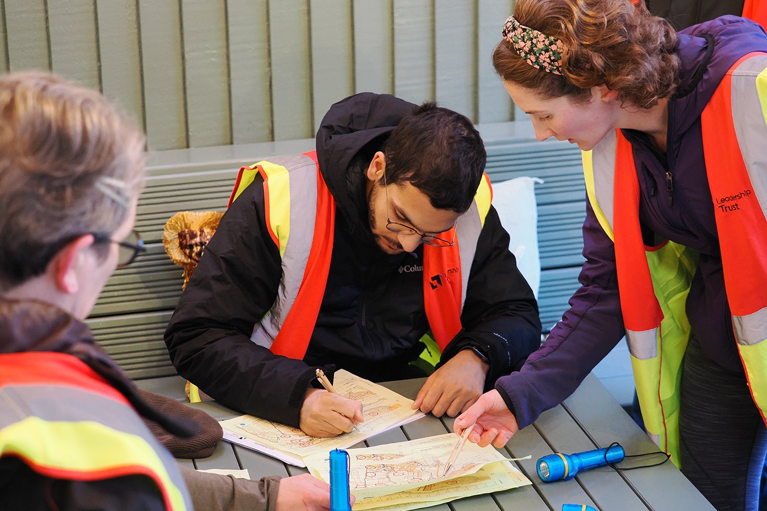 Group of four people wearing safety vests, sitting and standing at a table, collaborating on drawings or plans, with flashlights and pens on the table.