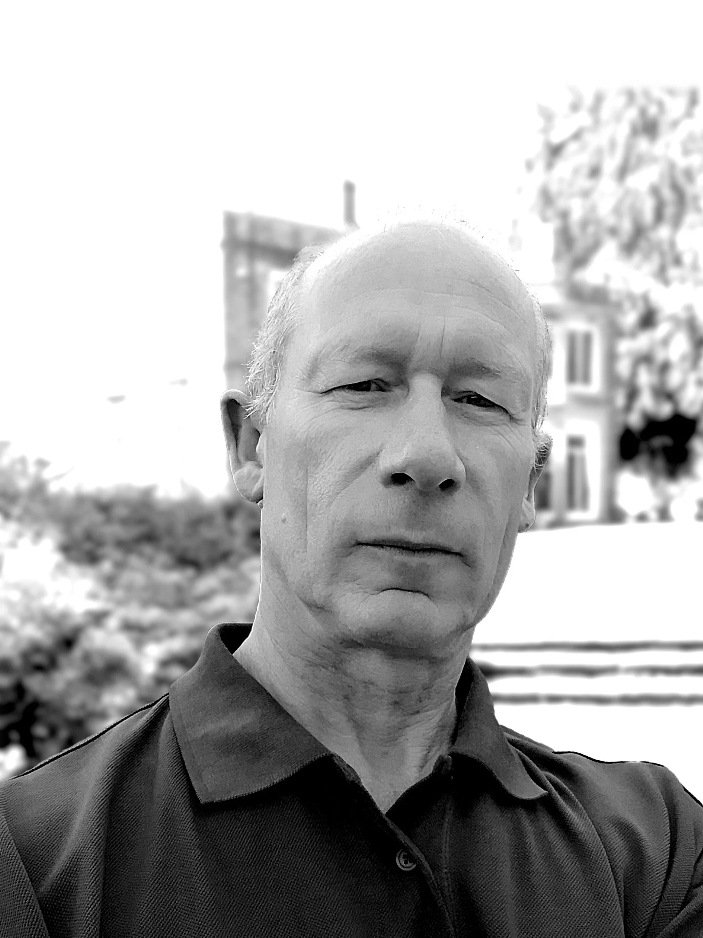 Black and white close-up of an older man with light skin, short hair, and a serious expression, wearing a collared shirt outdoors with trees and a building in the background.