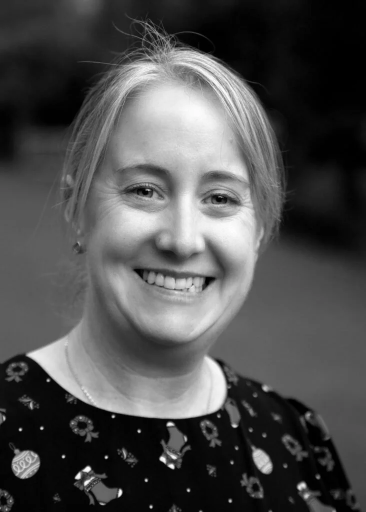 A smiling woman with blonde hair, wearing a patterned top, outdoors in black and white.