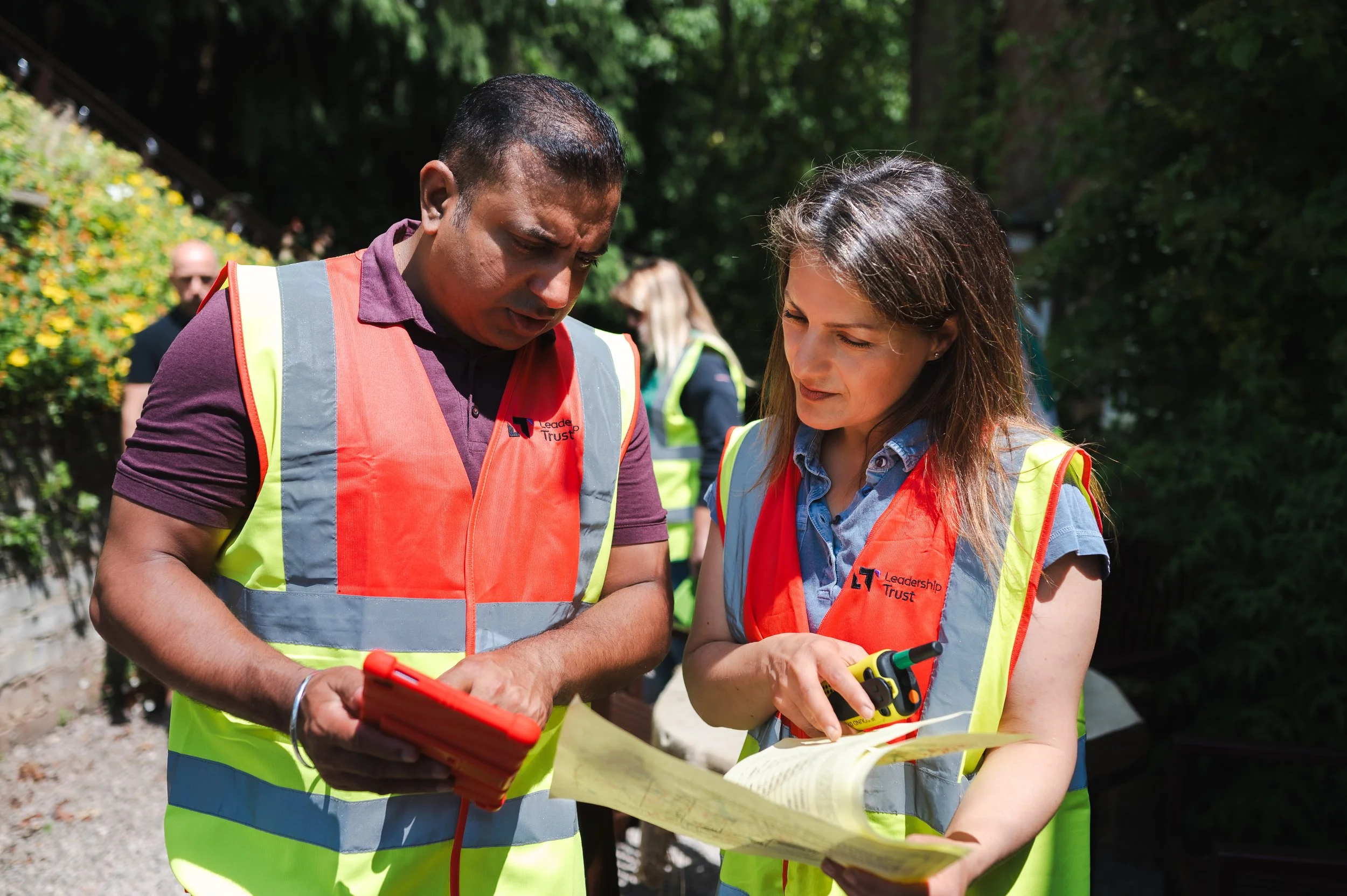 Two people wearing high-visibility vests consulting a map outdoors with greenery in the background.
