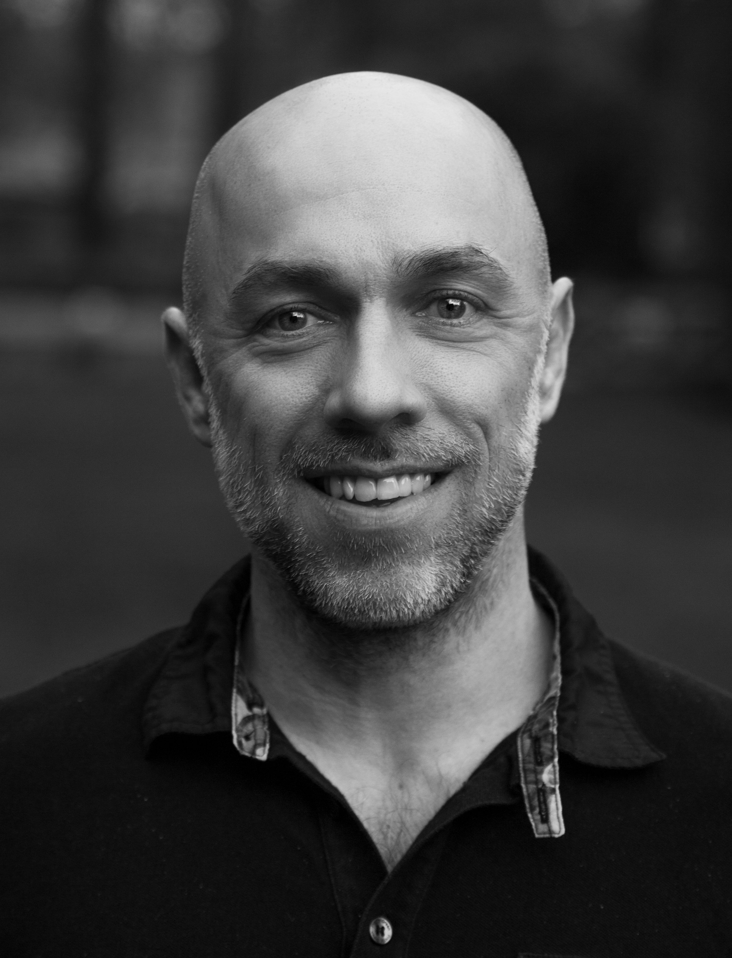 Black and white portrait of a smiling man with a bald head, light facial hair, wearing a dark shirt with a patterned collar and a jacket, standing outdoors.