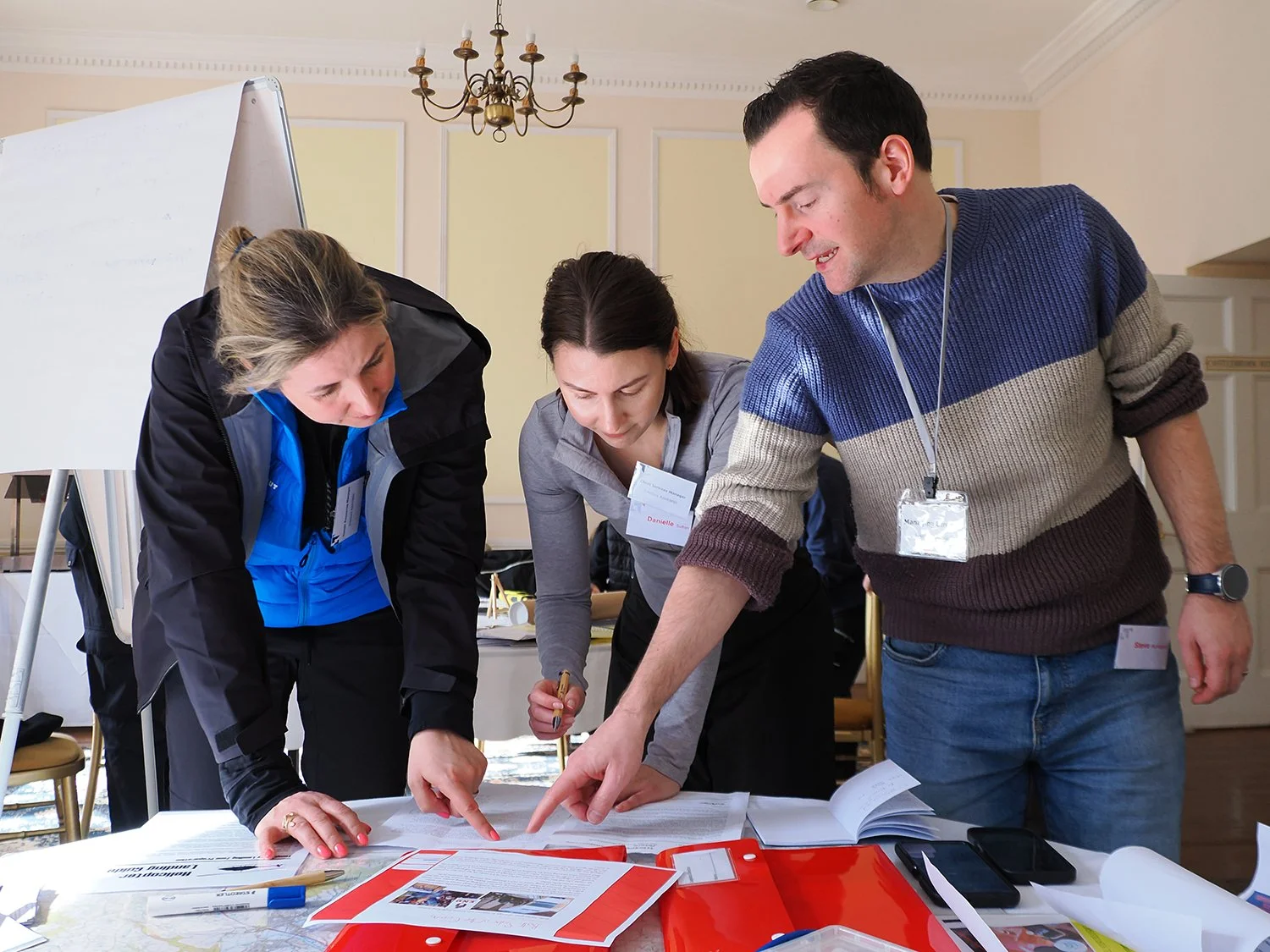 Three people standing around a table, examining documents and papers, in a room with classical decor and a chandelier.