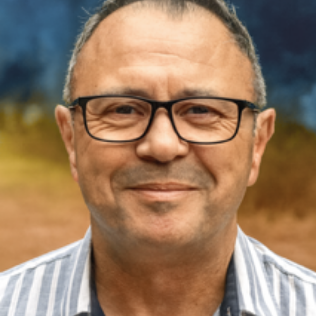 Close-up of a smiling man with glasses and short gray hair, wearing a collared shirt, outdoors with blurred fall foliage in the background.