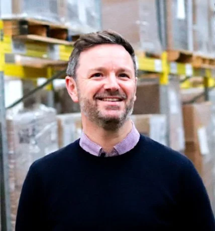 A man with short dark hair and a beard smiling in a warehouse setting, wearing a dark sweater over a collared shirt.