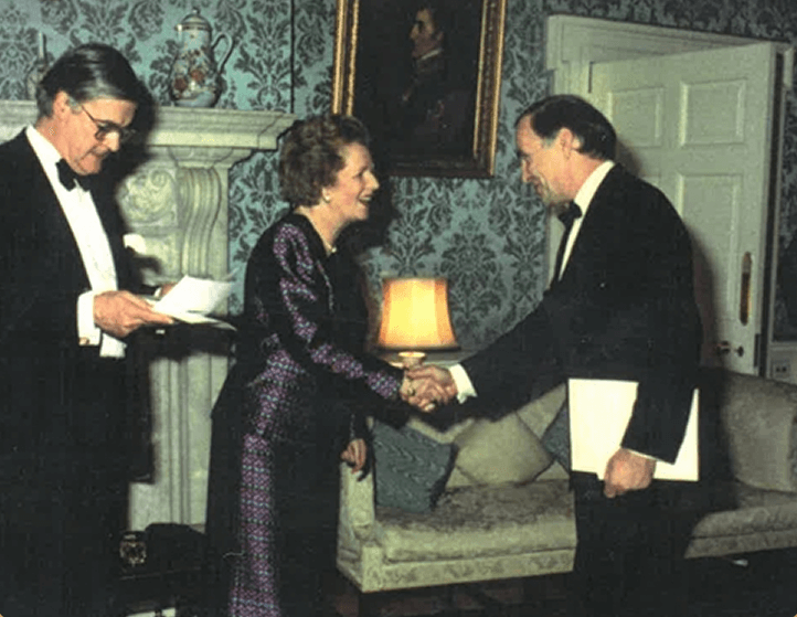 A woman and a man in formal attire shaking hands in a warmly decorated living room, with a man to the side reading papers, and a portrait hanging above a fireplace.