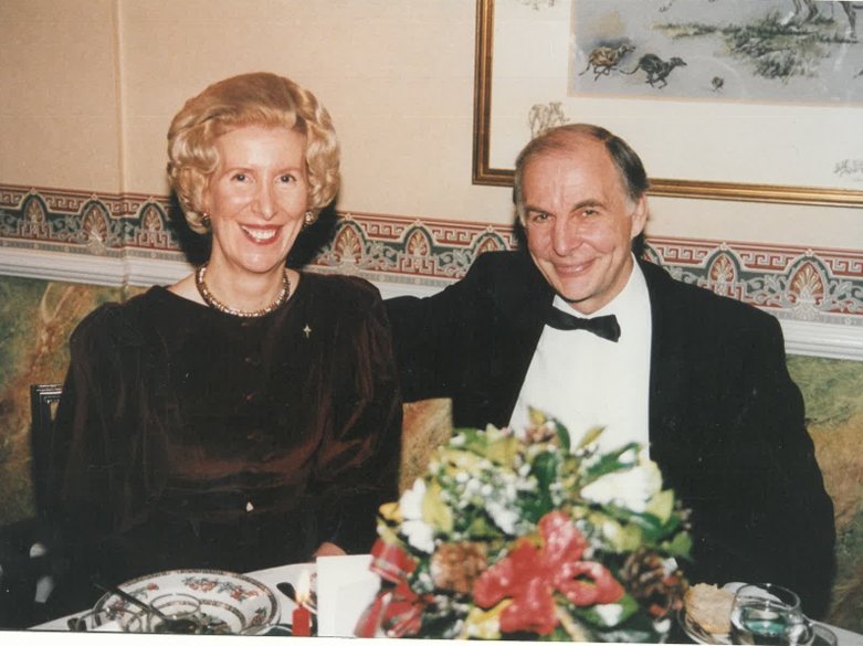 A man and woman dressed in formal attire sitting at a dinner table with a floral centerpiece, in a room with decorative wallpaper and a framed painting on the wall.
