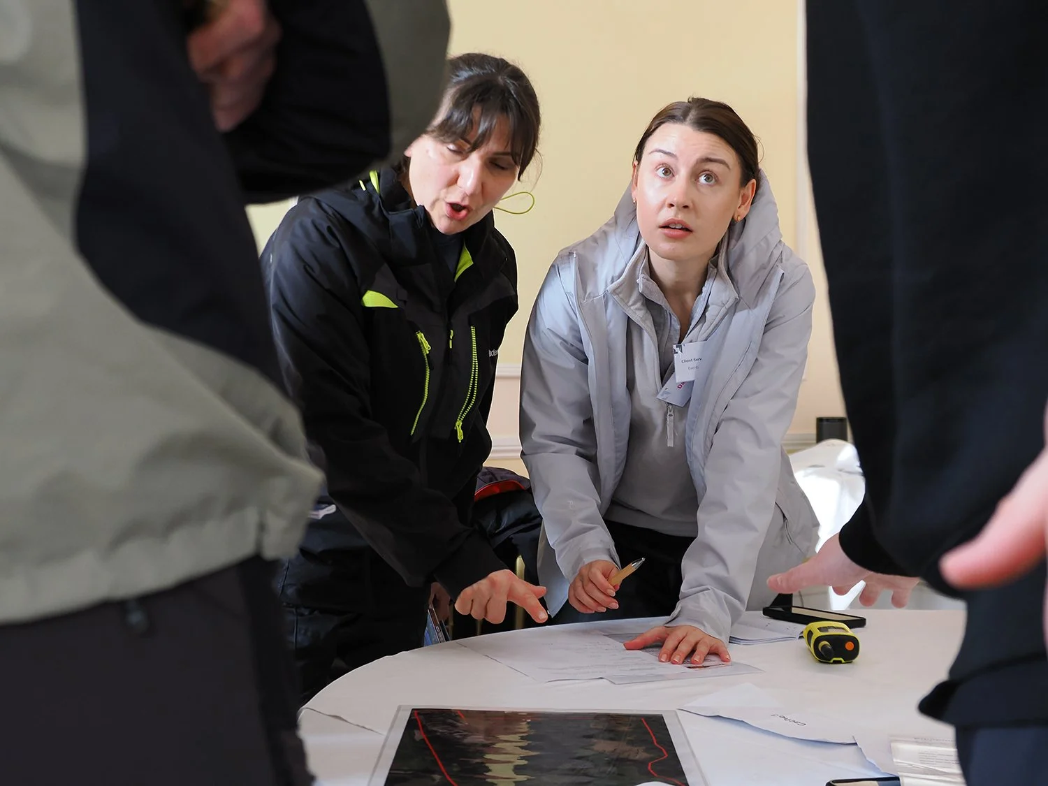 Two women and two men gathered around a table, looking concerned and pointing at a document and a map. The women are wearing white and black jackets, while the men are partially visible. They appear to be discussing an emergency or situation.