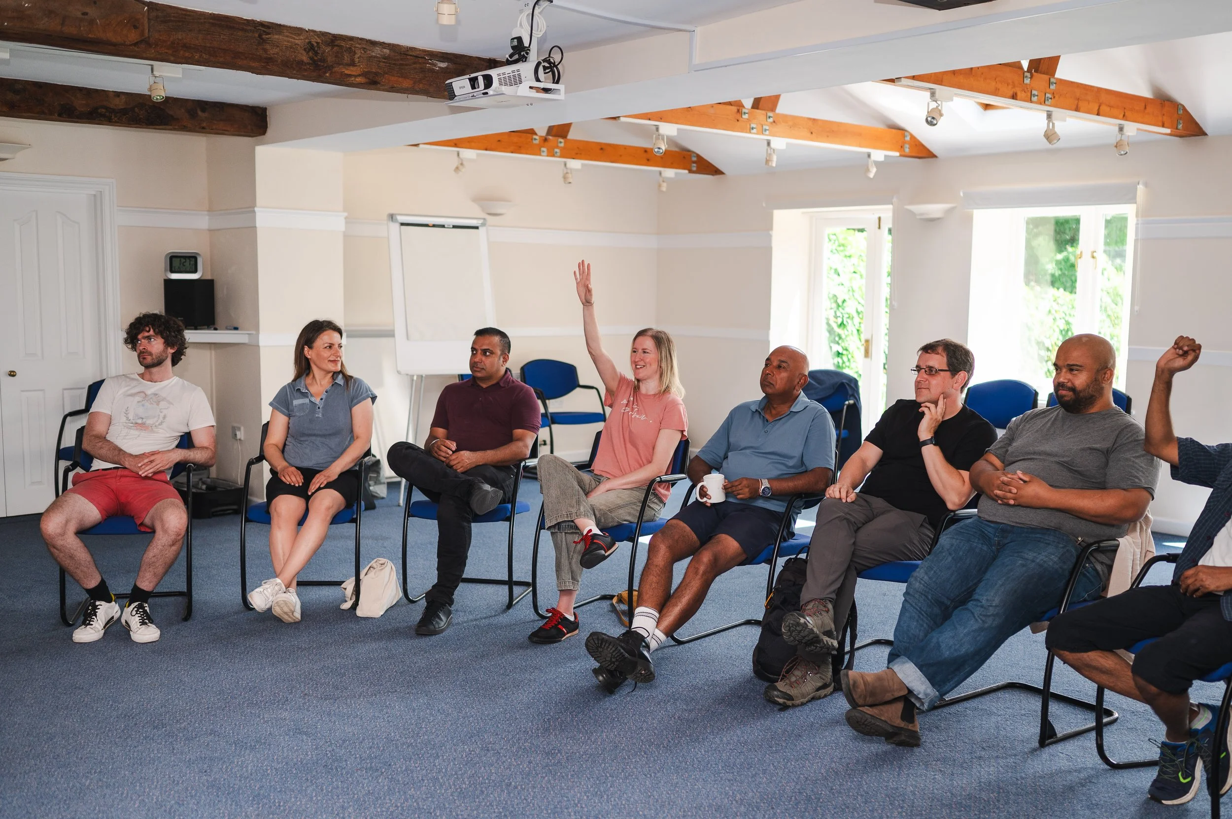A diverse group of seven people seated in chairs in a bright conference room, one woman with blonde hair is raising her hand, others are listening attentively, some holding cups, with windows letting in natural light and a ceiling-mounted projector.