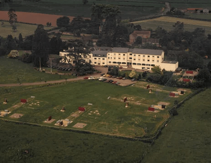 Aerial view of a large multi-story white building surrounded by a parking lot and green fields with small red-roofed structures.