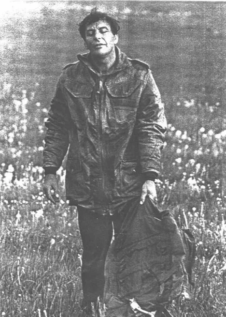 A man standing outdoors in a grassy field holding a backpack, wearing a heavy jacket with a serious expression.