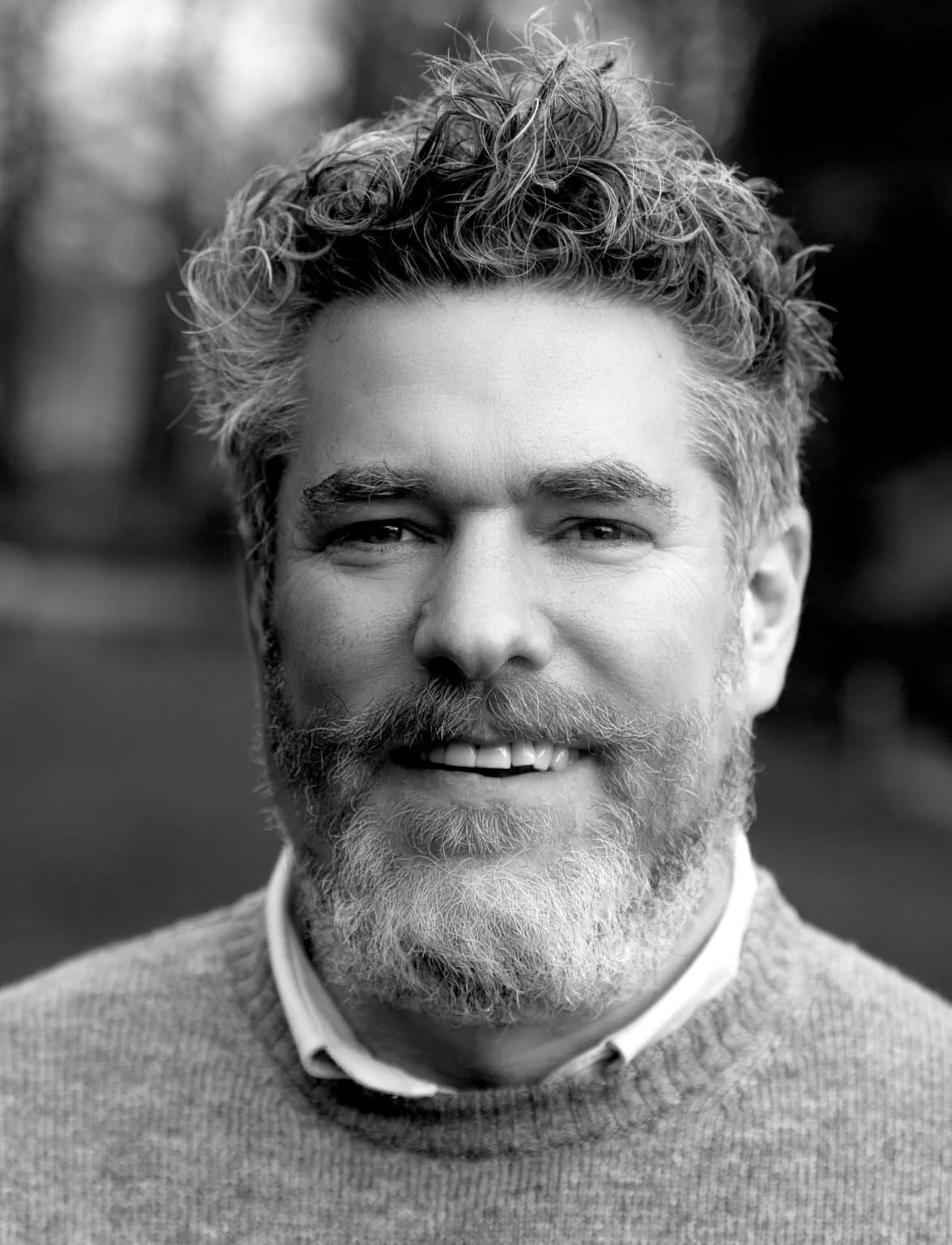 Black and white portrait of a smiling middle-aged man with curly hair, a full beard, and wearing a sweater over a collared shirt.