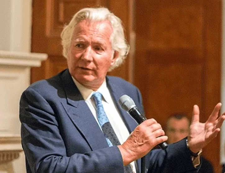 An older man with white hair, wearing a dark blue suit and a white shirt with a blue tie, is holding a microphone and speaking. He is gesturing with his left hand while standing in front of a wooden background.
