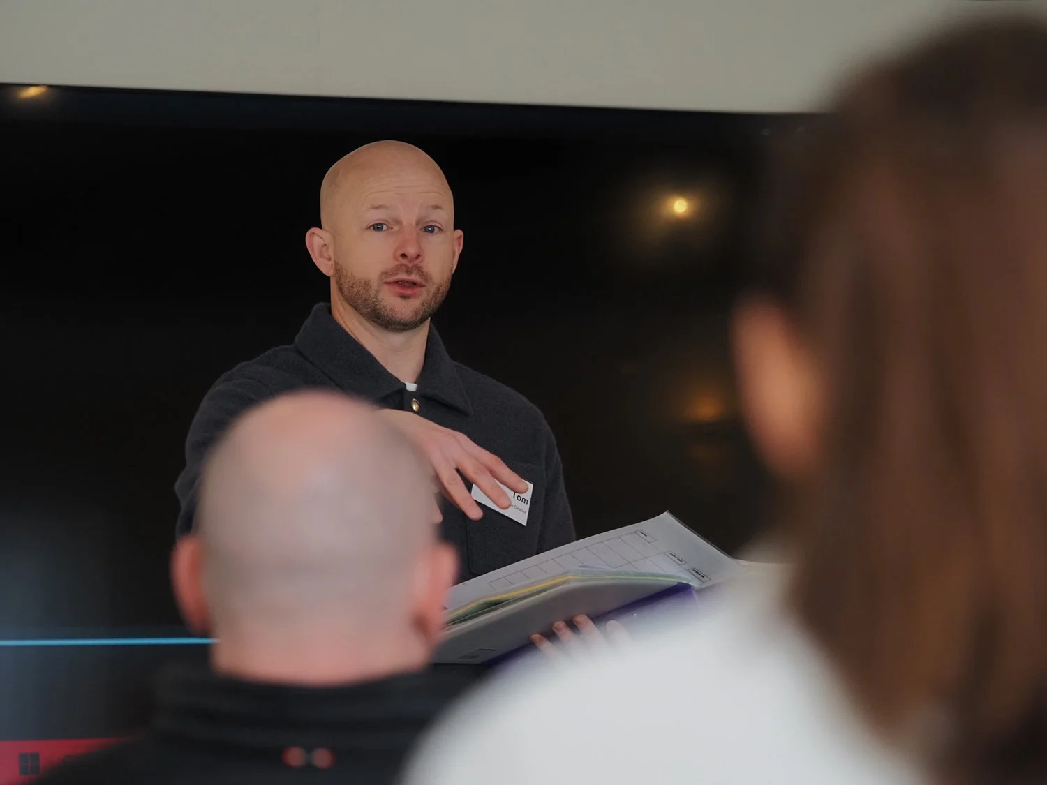 A man with a bald head and beard, wearing a dark shirt, is speaking to an audience. He has a name tag that says 'Tom'. He is holding a folder or clipboard in his hand. The audience is partially visible in the foreground, with the back of two heads visible.