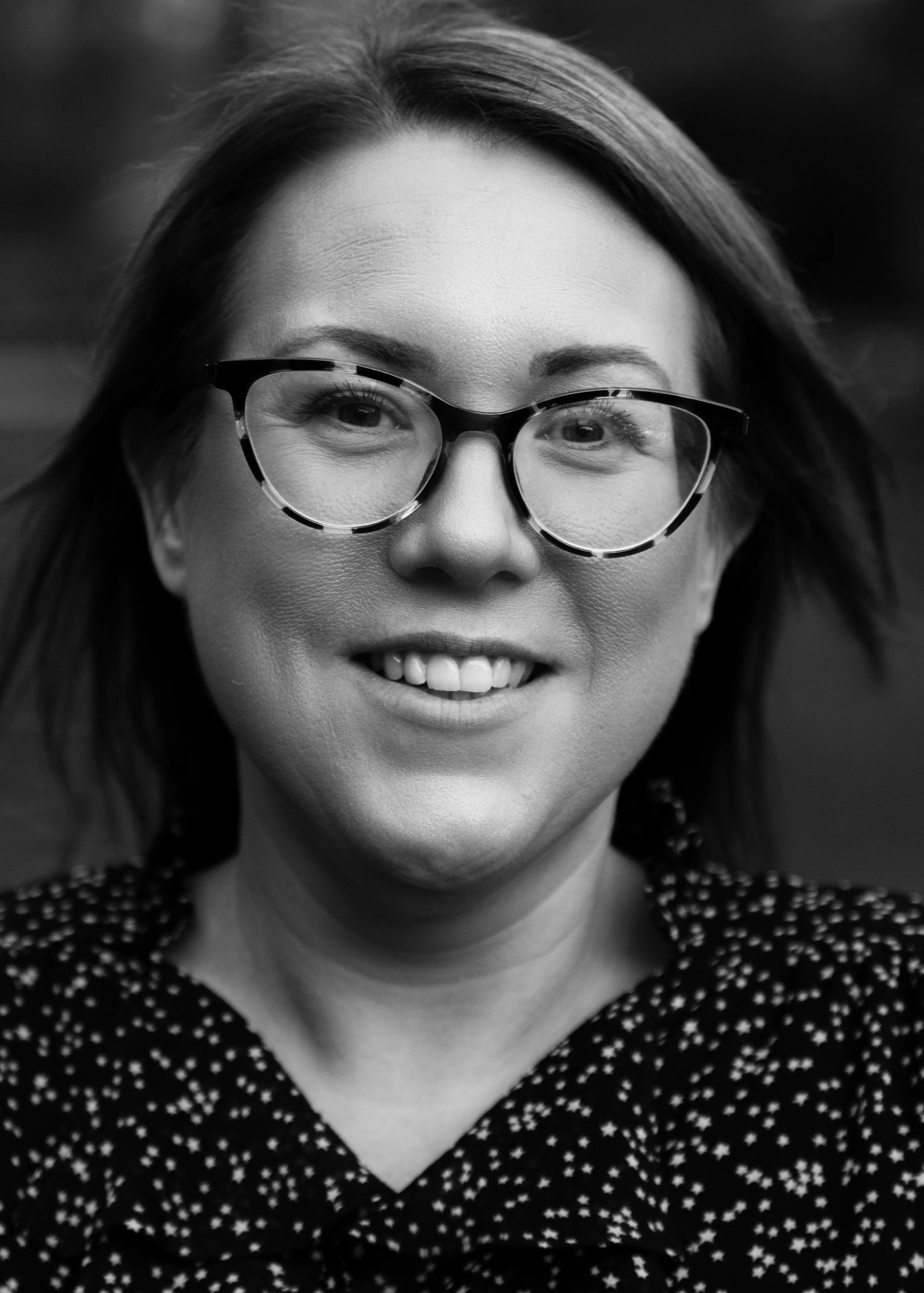 Close-up black and white portrait of a woman wearing round glasses and a polka-dotted top, smiling slightly.