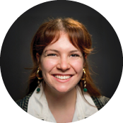 Smiling woman with reddish hair and earrings, wearing a white shirt and denim jacket, against a dark background