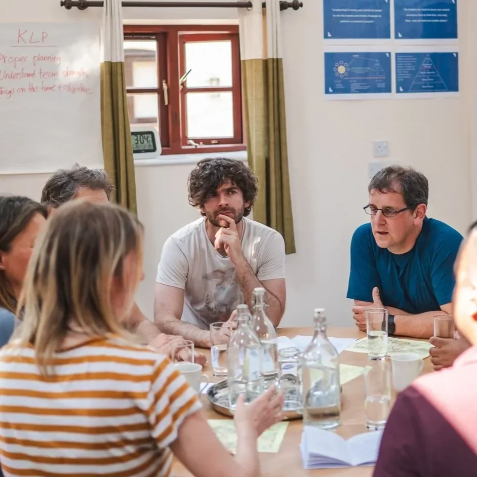 Group of six people sitting around a table in a meeting room, with bottles of water, glasses, and papers. Two people are speaking; one woman in a striped shirt and one man in a blue shirt. Three others are listening; one man with curly hair and beard, and two women partially visible. The room has white walls, a small window, and blue posters on the wall.