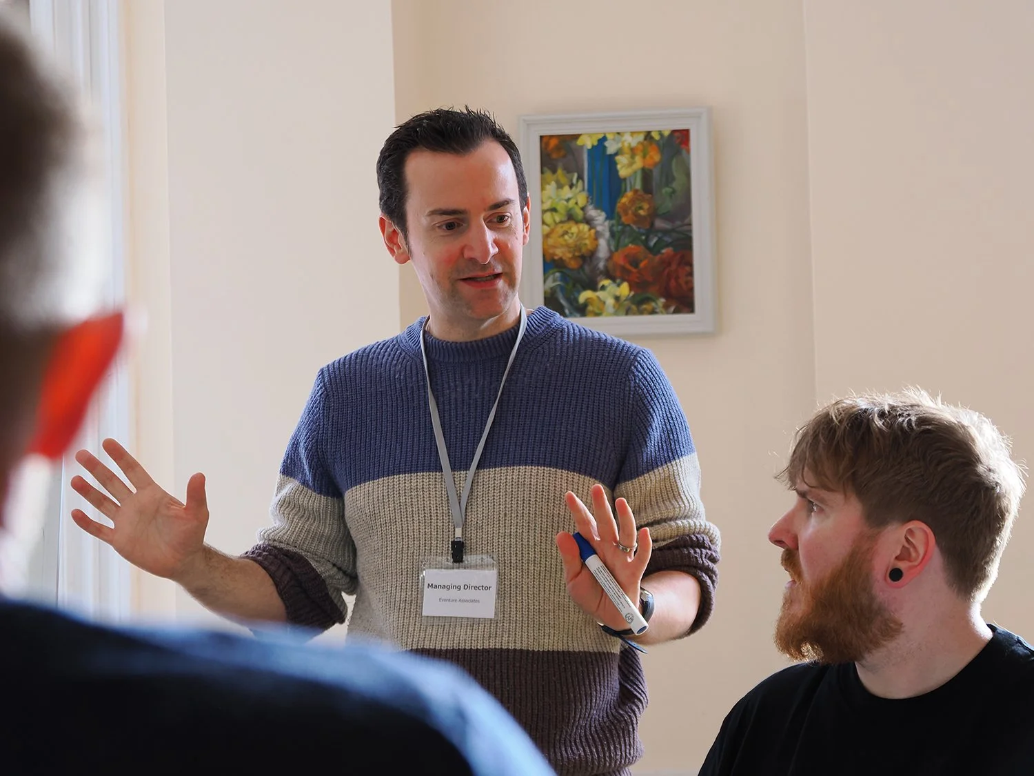 A man with a short dark hairstyle and a sweater, wearing a name badge, appears to be speaking to a group of people during a meeting or workshop. He is holding a marker and gesturing with his hands. A man with a beard and earrings is listening, seated at the table. There is a painting of flowers on the wall in the background.