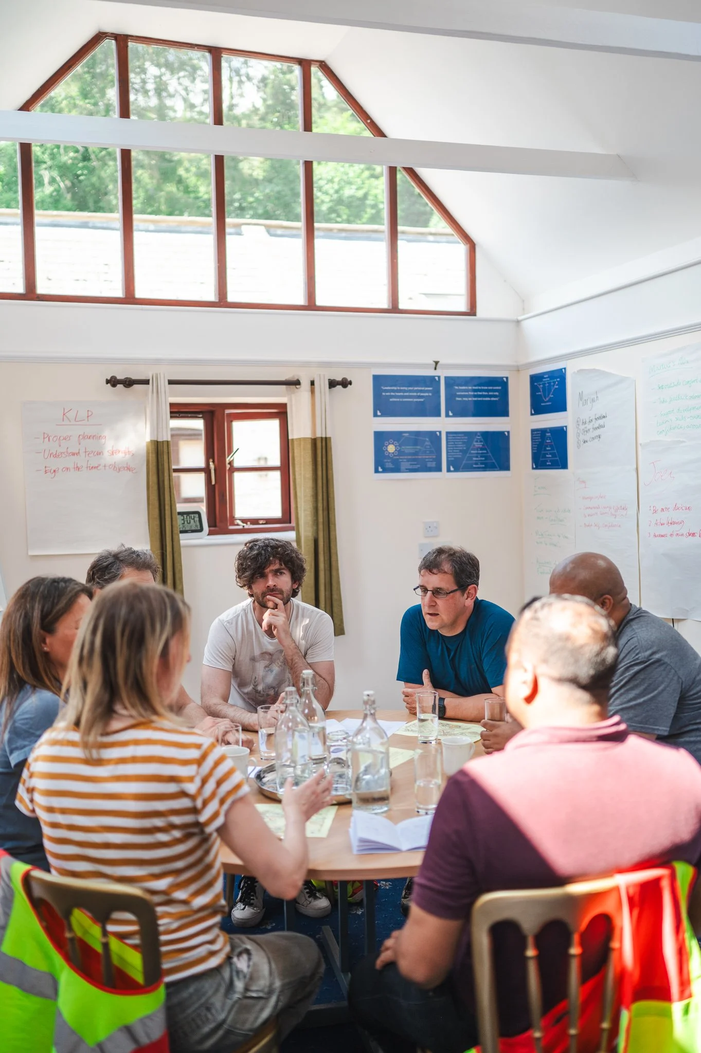 A group of people sitting around a table in a bright room with large windows and white walls, engaged in a discussion or meeting.