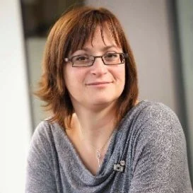 A woman with shoulder-length brown hair and glasses, wearing a gray top, smiling in an indoor setting.