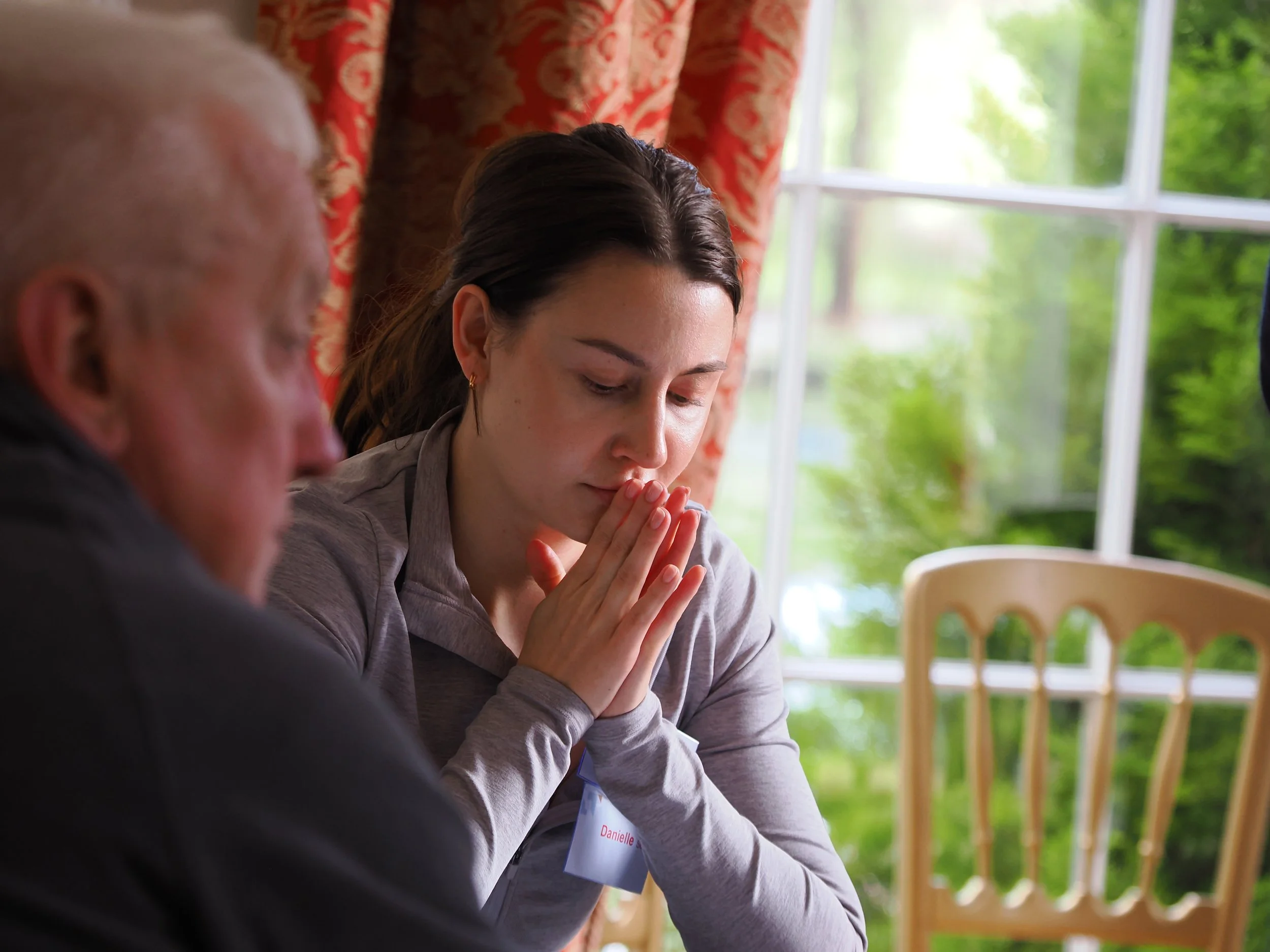 A woman with dark hair praying with her eyes closed and hands clasped, sitting at a table in a room with large windows and green trees outside.