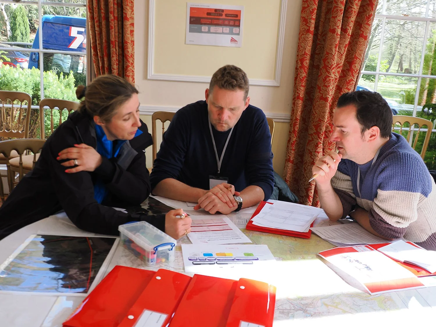 Three people sitting at a table in a room with large windows and red patterned curtains, engaged in a discussion with documents and red folders on the table.
