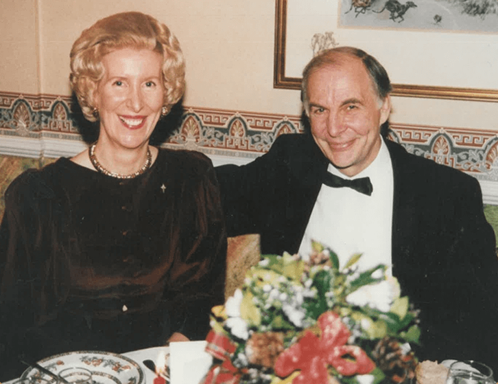 A woman and a man dressed in formal attire sitting at a dinner table with a flower centerpiece, smiling at the camera.