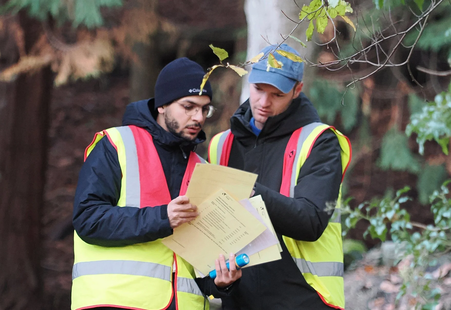 Two men wearing black jackets, glasses, and yellow safety vests with reflective stripes are standing outdoors, examining documents together. One man holds a rolled-up item and the other is pointing at the papers. They are surrounded by trees and foliage.
