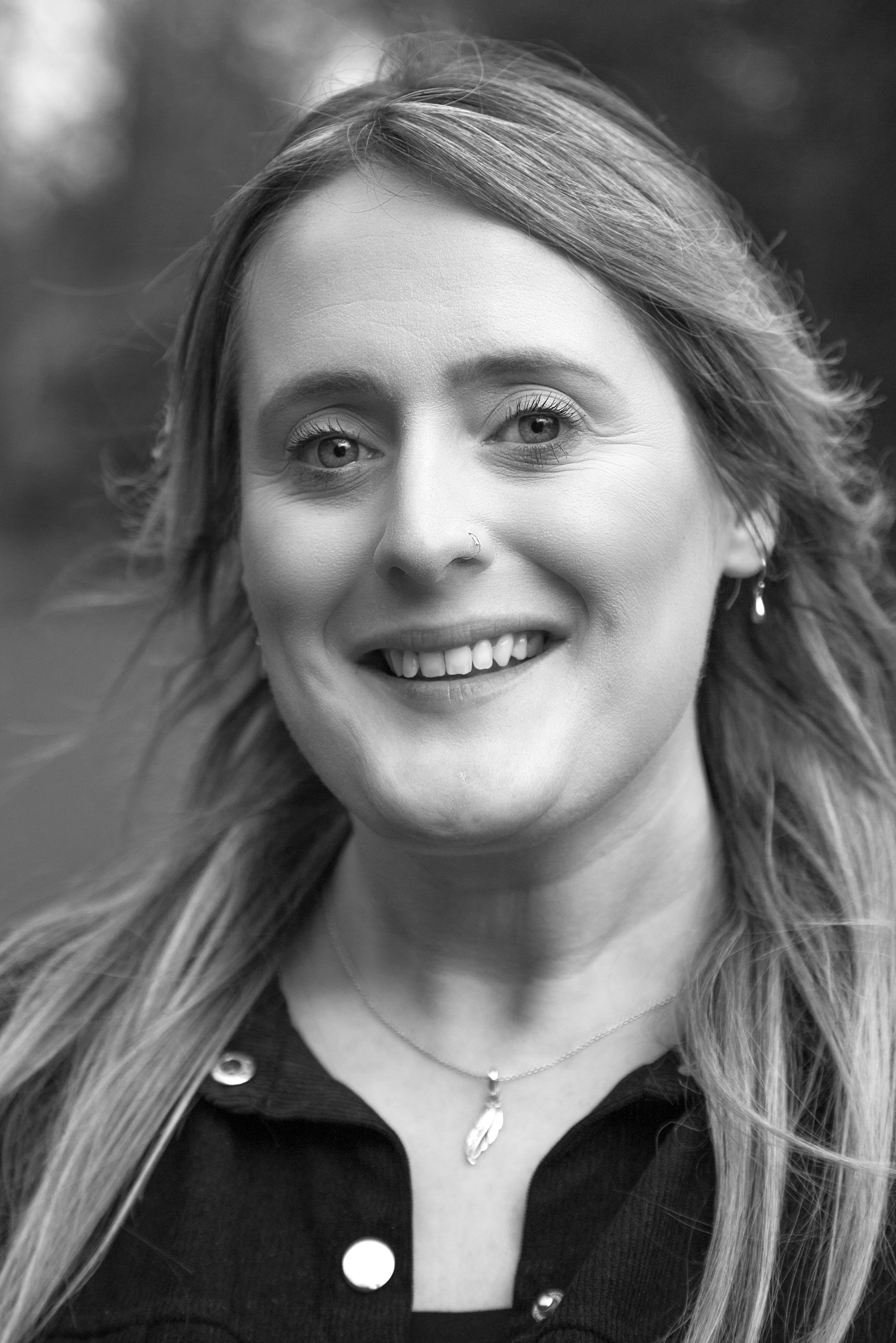 Close-up black and white photo of a woman smiling with long hair, wearing jewelry including earrings, a necklace with a feather pendant, and a shirt with large buttons.