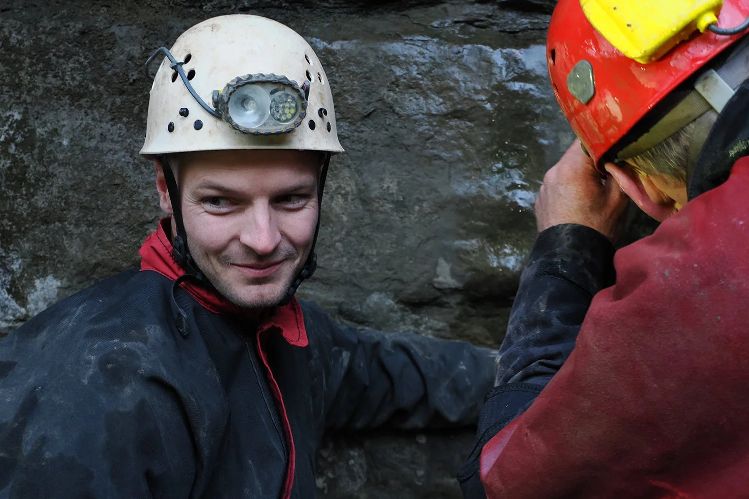 Two men in helmets and outdoor gear, engaged in caving or spelunking inside a rocky cave.