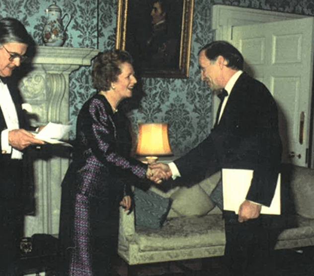 A woman and a man are shaking hands in a formal setting with a woman holding a notepad nearby. The room has patterned wallpaper, a fireplace with a decorative item on it, a lamp, and a portrait hanging on the wall.
