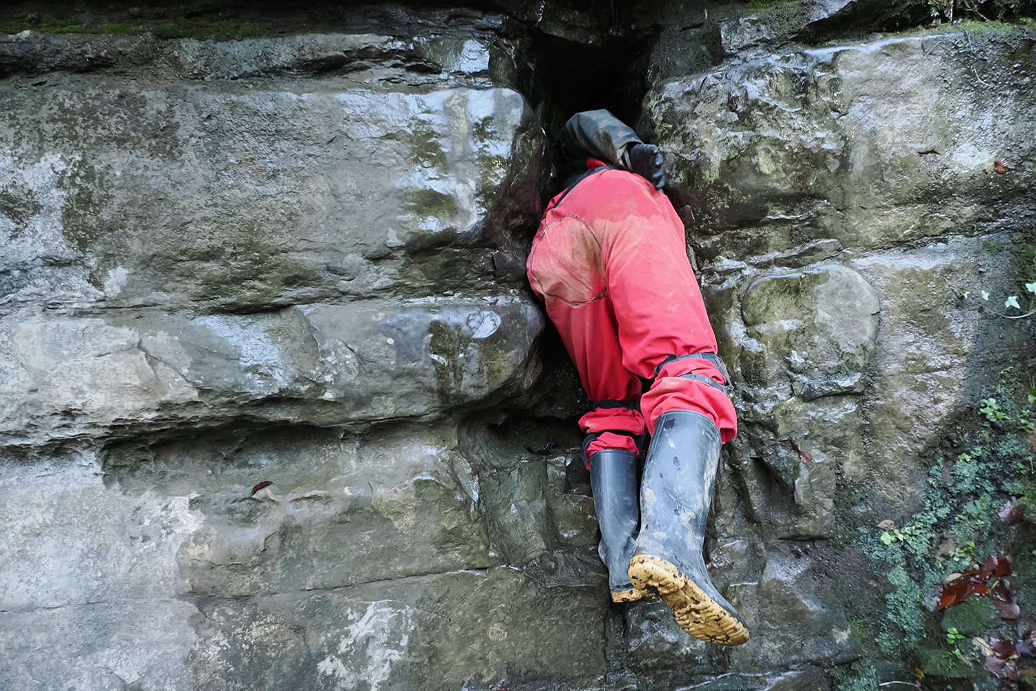 Person wearing red pants and black rain boots climbing a wet rock face outdoors.