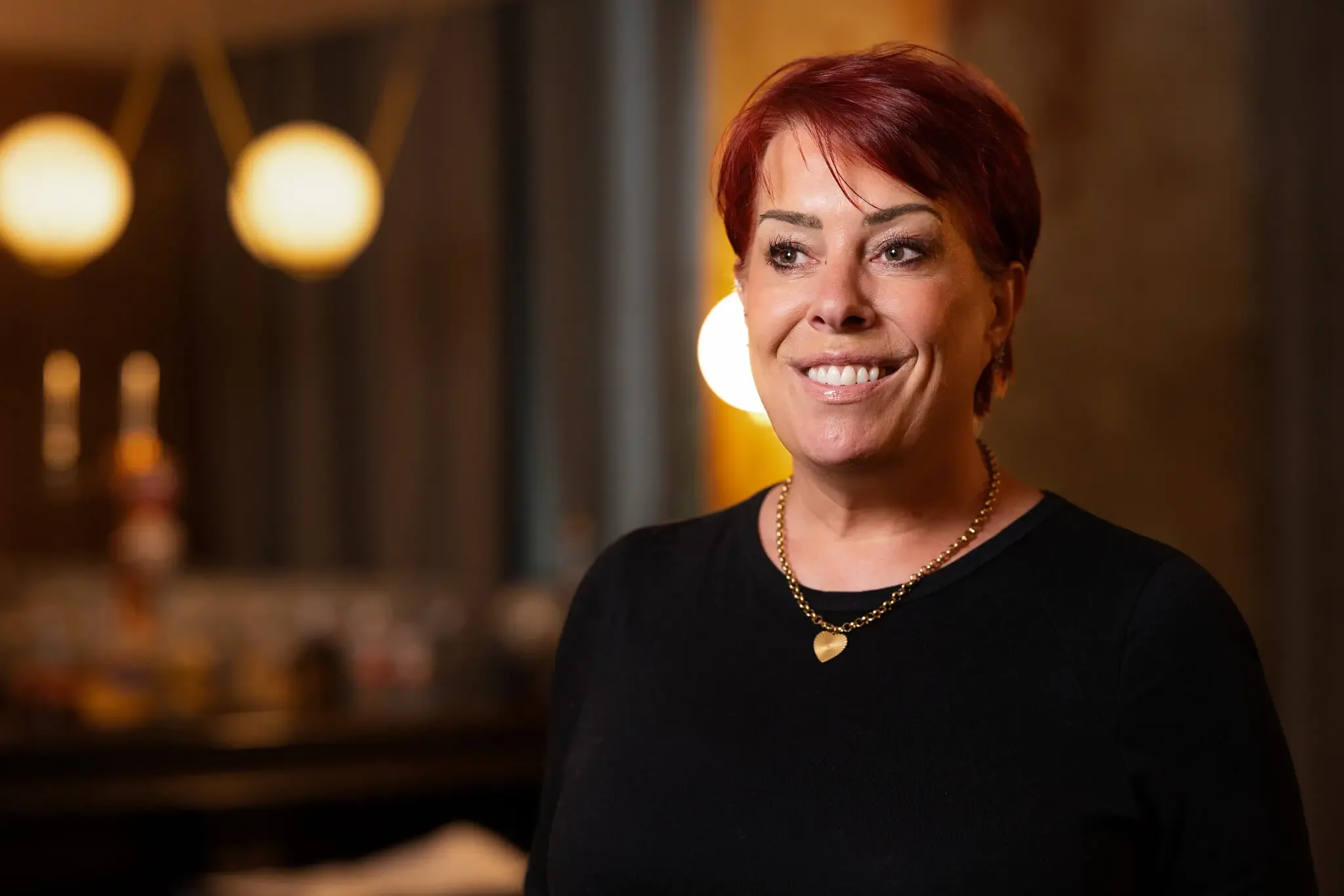 A woman with short red hair smiling, wearing a black top and a gold heart-shaped necklace, standing indoors with warm lighting and blurry background.