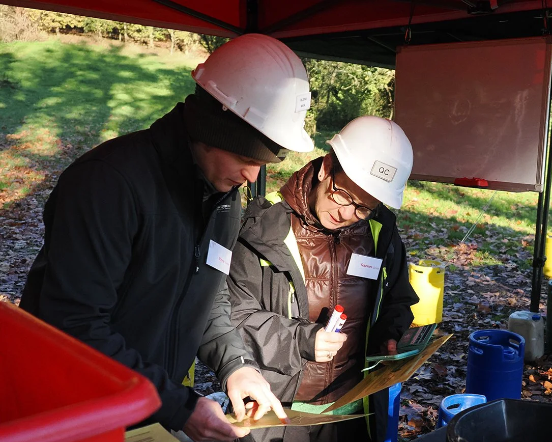 Two individuals wearing white safety helmets and dark jackets examining documents outdoors under a red canopy, with trees and fallen leaves in the background.